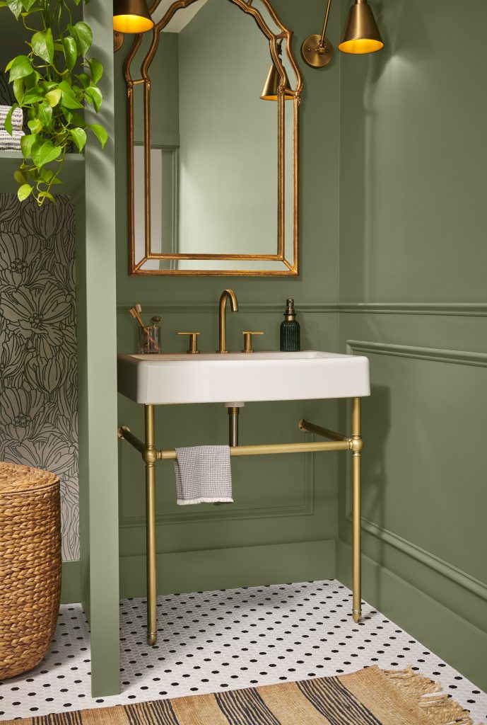 A small powder room with walls painted in “Ecological,” featuring a white pedestal sink with brass fixtures, a gold-framed mirror, and warm, modern accents.