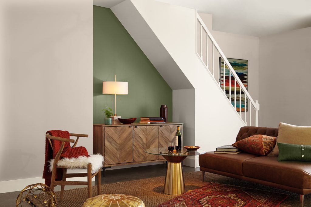 A basement living area painted with an earthy green paint called  “Ecological” as the accent wall under the staircase, styled with a wood sideboard, leather sofa, and warm, layered decor.