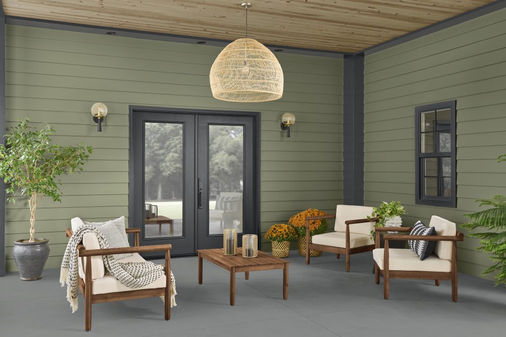 A covered patio with green siding, black French doors, wood ceiling, woven pendant light, and outdoor seating.