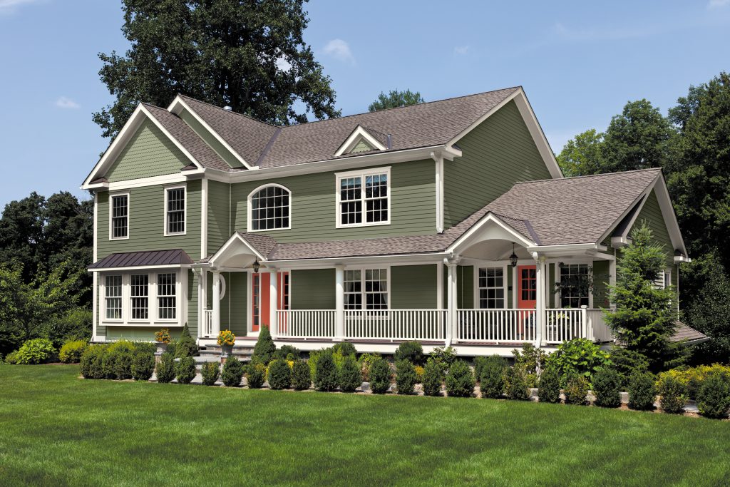 A large dight green two-story home with white trim, wraparound porch, red front doors, and manicured lawn.