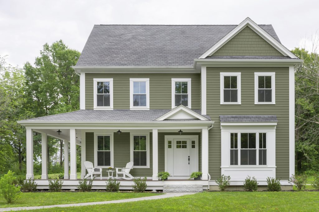 A two-story green colored farmhouse-style home with white trim, a wraparound porch, and a green front lawn.