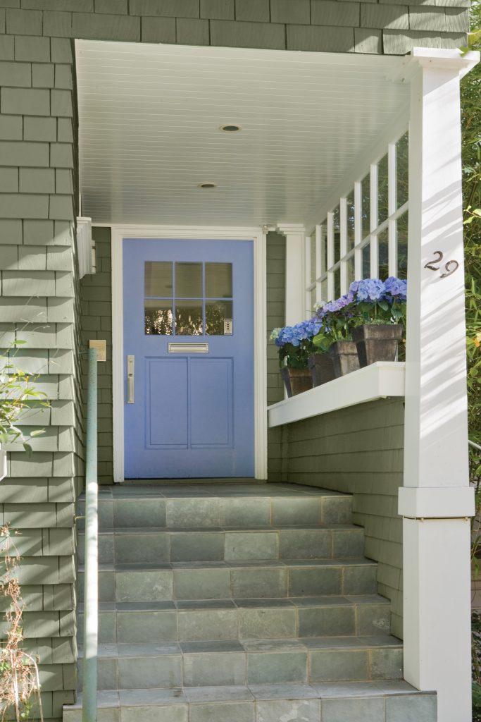 A green shingle house entryway with a blue front door, slate steps, white trim, and potted blue flowers.