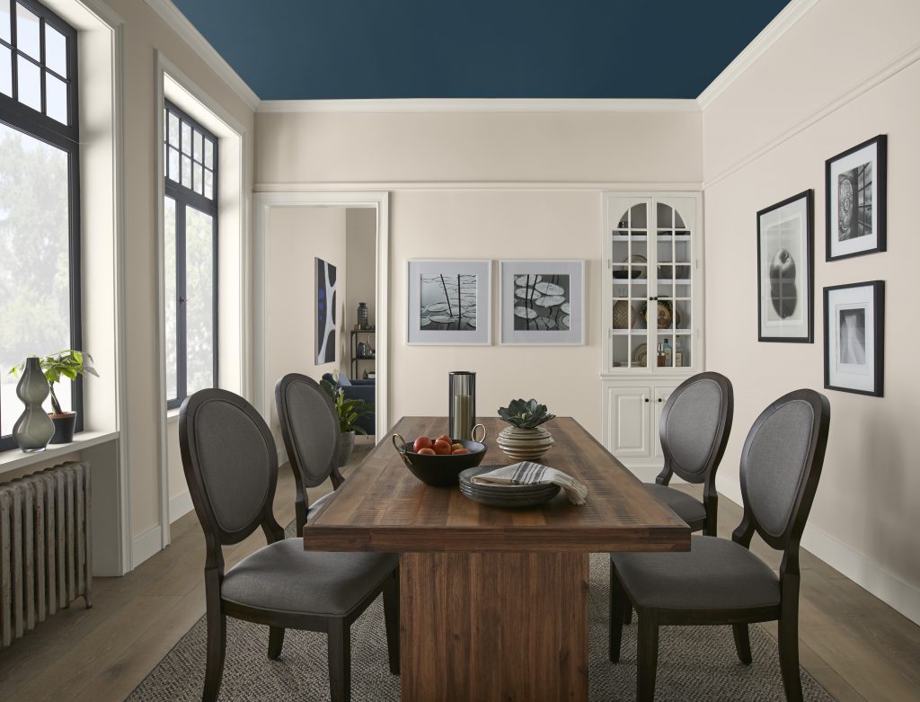 A dining room with Wheat Bread walls, crisp white trim and a dark blue ceiling, featuring a wood dining table, gray upholstered chairs, tall black-framed windows and framed wall art.