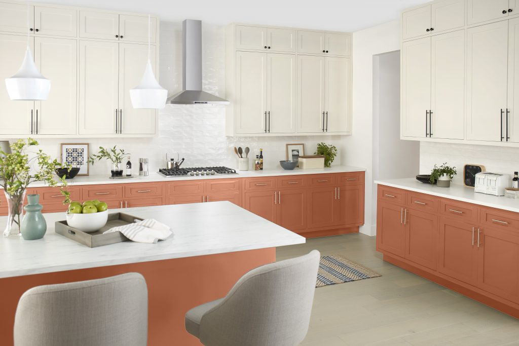 A bright kitchen with warm terracotta paint color on the lower cabinets, creamy upper cabinets, white countertops, and modern pendant lighting.