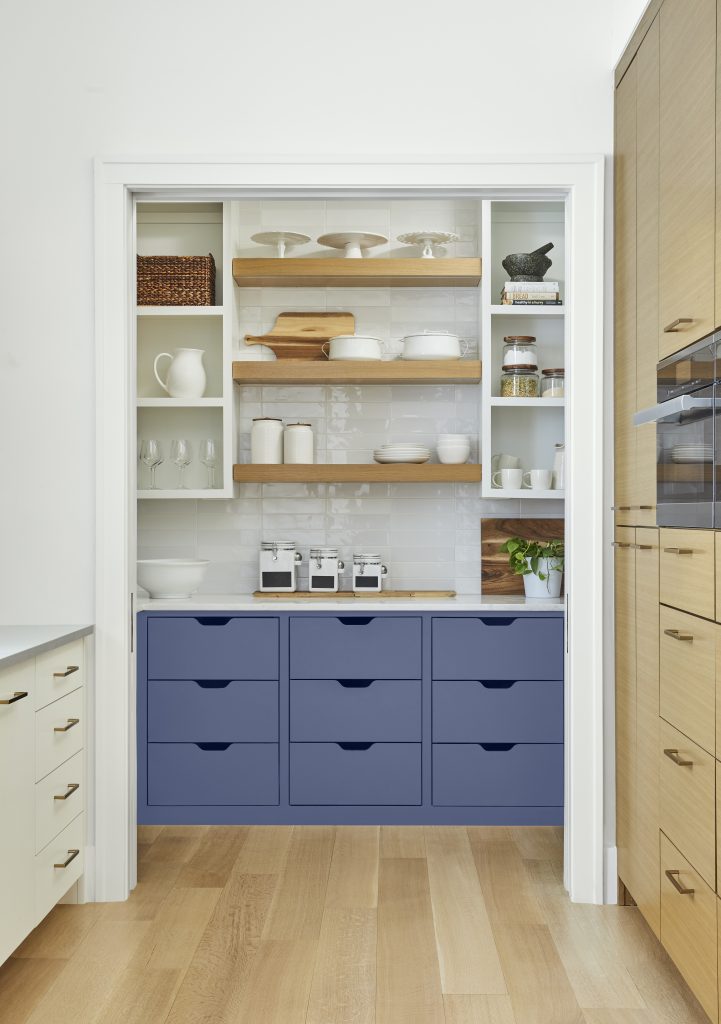 A pantry nook with blue-purple lower cabinets that create a surprise pop of color, paired with open wood shelving, white built-in storage, natural wood cabinetry, and light wood floors.
