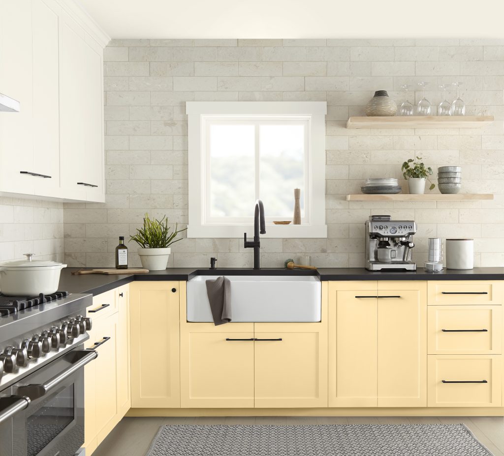 A bright kitchen with soft yellow paint color on the lower cabinets, a farmhouse sink, black hardware, and a light stone backsplash. The soft yellow cabinetry helps the small kitchen feel larger, brighter, and more inviting by reflecting light and creating a warm, airy atmosphere.