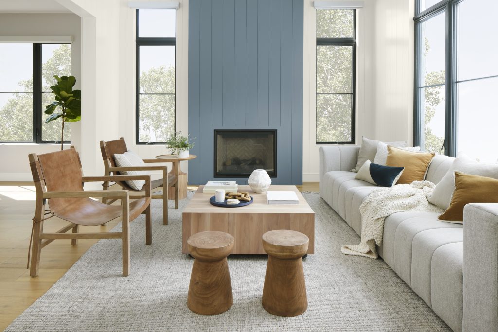 A light-filled living room with a blue painted fireplace wall, white sectional sofa, natural wood chairs, chunky wood stools, large windows, and layered neutral textures.