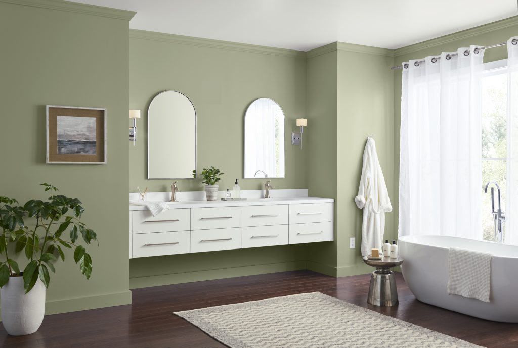 A spa-inspired bathroom with soft sage green walls, a double white vanity with arched mirrors, freestanding soaking tub, sheer white curtains, dark wood floors, and layered neutral textures.