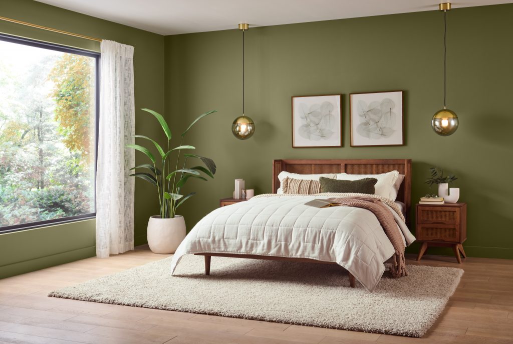 A Mid-century modern bedroom with olive green walls, a wood bed frame, woven nightstands, globe pendant lights, neutral bedding, a textured rug, and large window overlooking greenery.