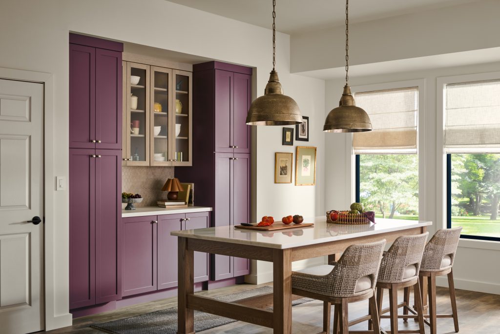 A rich and inviting kitchen nook with purple-red paint on the cabinetry, wooden table, woven chairs, brass pendant lights, and large windows.