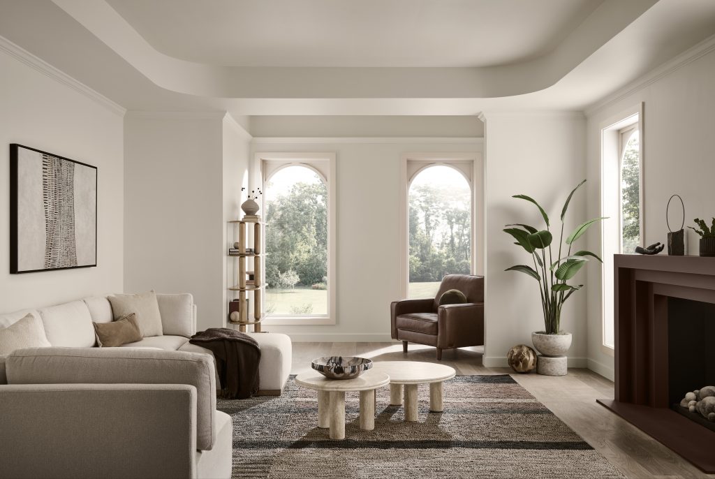 A living room showcasing several neutral paint colors for living room. The walls were painted in Wheat Bread and this room is featuring arched windows, a light neutral sofa, layered neutral textures, warm wood accents and a fireplace. 