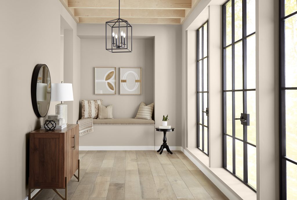 A light-filled hallway with walls painted in Wheat Bread, creating a warm neutral backdrop for black-framed windows, pale wood flooring, exposed ceiling beams and a cozy built-in seating nook.
