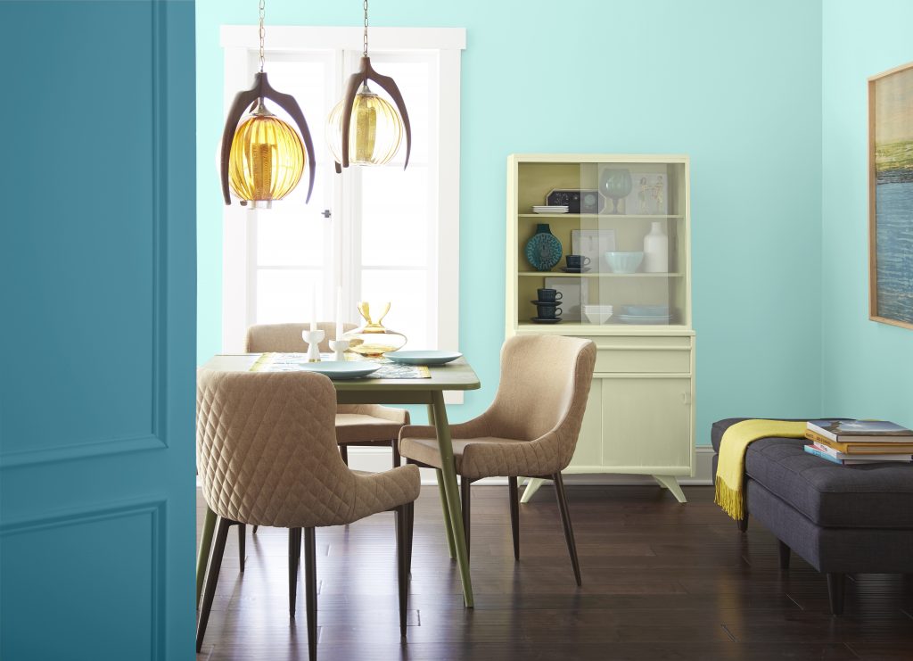 A bright dining nook with aqua blue walls, retro pendant lights, upholstered chairs, dark wood floors, a light chartreuse china cabinet, and a sitting area with colorful accents.
