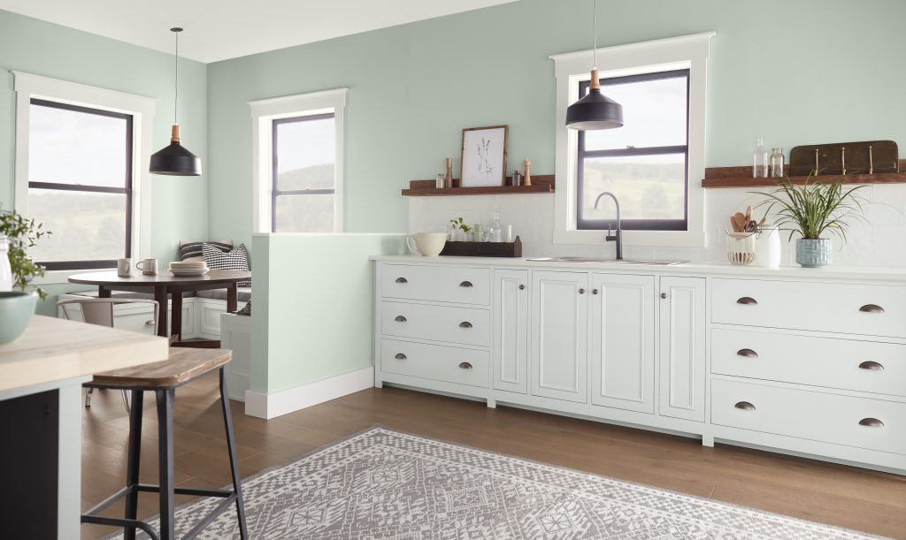 A bright kitchen with soft green walls, white cabinetry, black pendant lights, wood shelving, and large windows. The green color gives the kitchen a fresh, calming feel while adding subtle contrast to the white cabinets and natural wood tones.