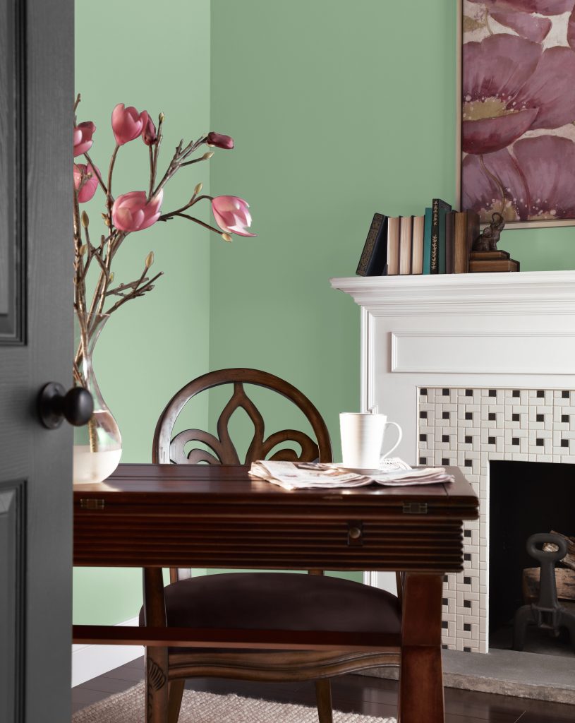 A Traditional study nook with soft green walls, a white fireplace, dark wood desk, vintage chair, pink flowering branches, and framed floral artwork for a refined, nature-inspired look.