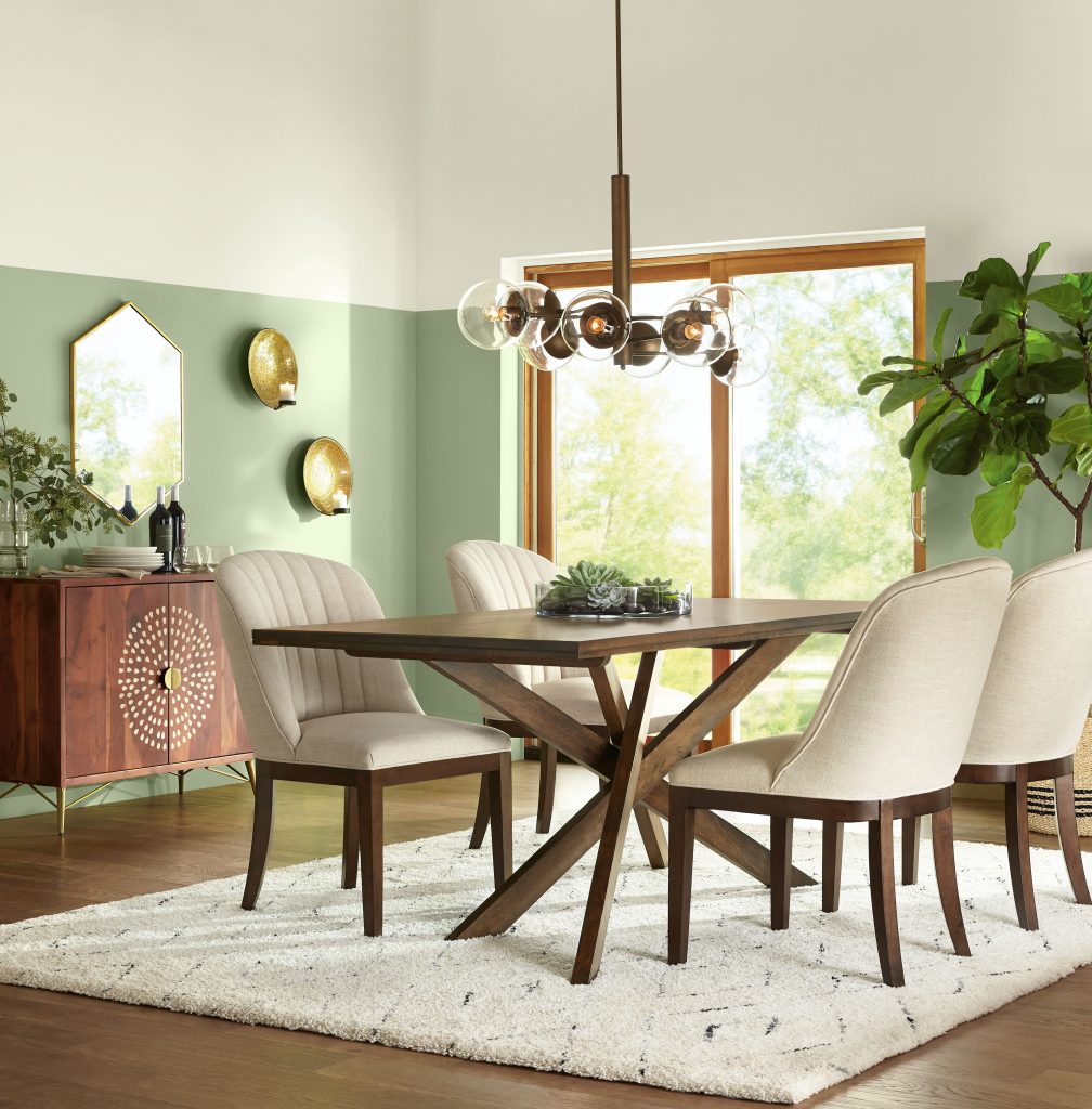 Dining room featuring Chopped Dill green wall paint with natural wood furniture, cream upholstered chairs, warm metallic accents, and botanical styling.