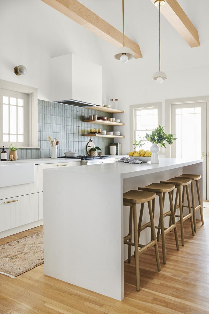 A layered, textured modern organic farmhouse kitchen with white walls and white cabinetry, a large light gray island, light wood beams, open shelving, wooden bar stools, pale blue tile on the backsplash, and warm natural wood floors.