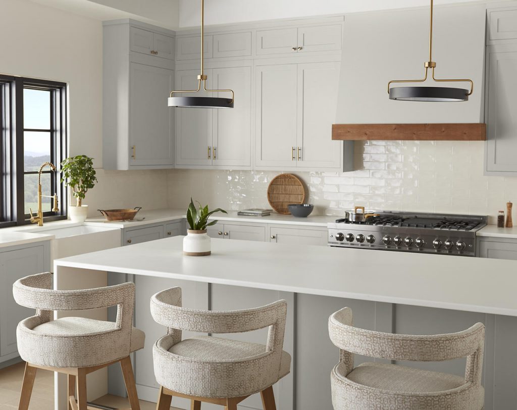 A white and light gray kitchen with warm natural wood range trim, brass hardware, brass pendant lights, white countertops, and textured neutral bar stools. The wood tones warm up the gray cabinetry, while brass hardware adds a modern, polished finish.