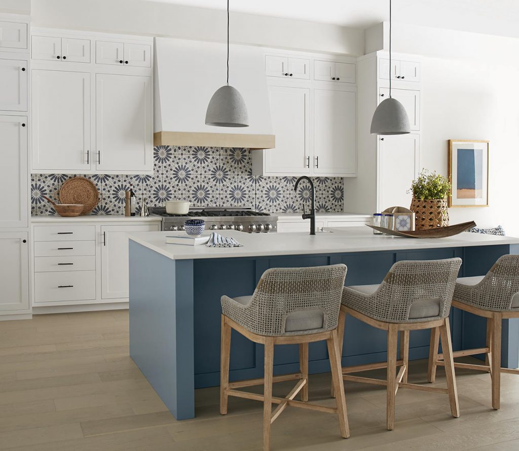 A white kitchen with a dusty and top-selling blue paint color on the island, blue patterned tile backsplash, white cabinets, gray pendant lights, woven bar stools, and light wood floors.