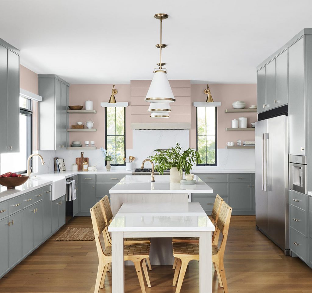 A bright kitchen with gray-blue cabinets, brass hardware, white countertops, a soft pink accent wall, wood dining chairs, and warm natural light. The gray-blue cabinetry brings a calm, sophisticated feel to the space, while the soft pink wall adds warmth and softness, creating a balanced kitchen that feels both fresh and inviting. Together, the colors add depth and character to this kitchen design.