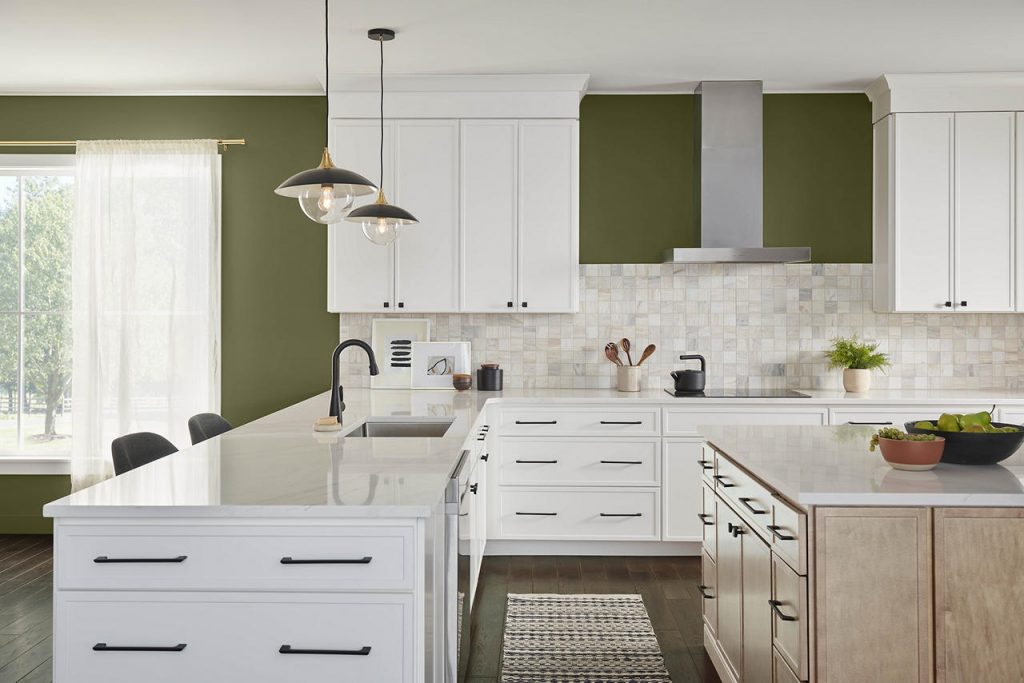 A large and expansive kitchen with olive green walls, white cabinetry, black hardware, light wood cabinetry, and white quartz countertops. The olive-green color adds natural warmth and earthy sophistication, creating a grounded, welcoming atmosphere. Paired with crisp white cabinets and light wood tones, it brings depth, contrast, and a timeless organic feel to the space.