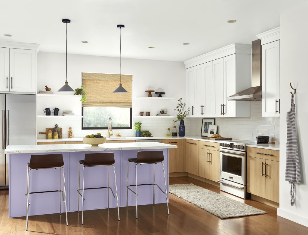 A kitchen with a lavender island, white upper cabinets, natural wood lower cabinets, white countertops, pendant lights, and warm wood floors. The soft purple shade makes the kitchen feel soft and approachable. Paired with crisp white countertops and natural wood cabinetry, this light purple feels fresh, balanced, and easy to use in everyday kitchen design.