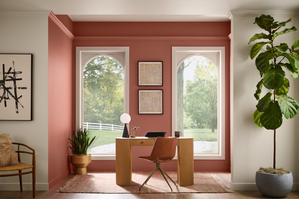 A home office nook with terra cotta painted walls, arched windows, a wood desk, leather chair, abstract artwork, woven accents, and a large leafy plant.