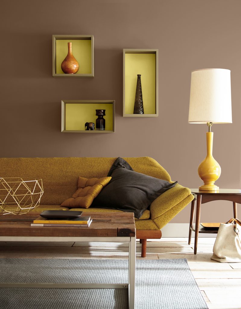 A Mid-century modern living room area with warm brown walls, a mustard yellow lounge chair, walnut coffee table, geometric decor, display shelves, and a sculptural table lamp creating a cozy retro atmosphere.