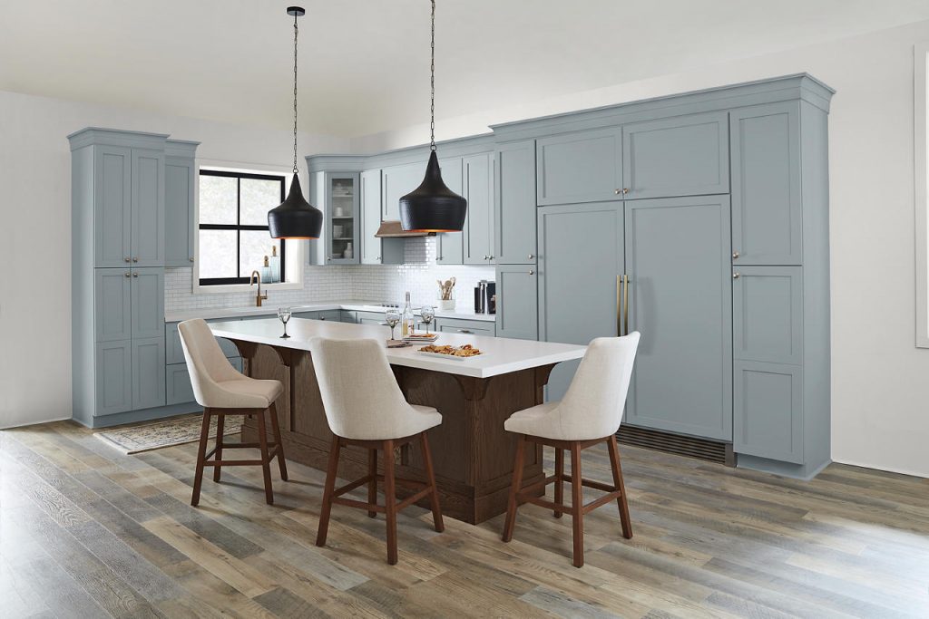 A kitchen with a popular soft blue paint color on the cabinetry, brushed brass hardware, a dark wood island, warm wood flooring, black pendant lights, and cream bar stools.