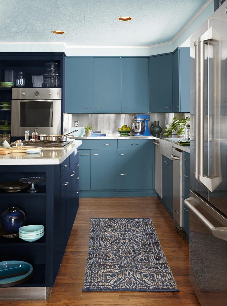 A blue color-drenched kitchen with deep navy island cabinetry, softer blue wall cabinets, and a lighter coodinating blue ceiling that creates a bold, immersive look. A blue patterned runner and blue dishware continue the layered palette, while stainless steel appliances and warm wood floors add balance and contrast.