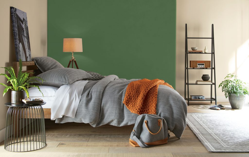 Bedroom with deep Garden Cucumber green accent wall, warm neutral walls, gray bedding, natural wood furniture, plants, and earthy orange accents.