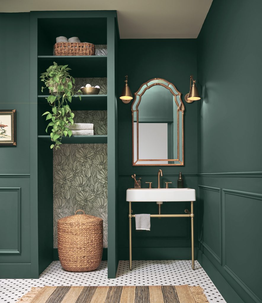 A moody bathroom with deep green walls and wainscoting, white sink with brass vanity, arched mirror, wall sconces, built-in shelving with plants and woven baskets, and a patterned tile floor.