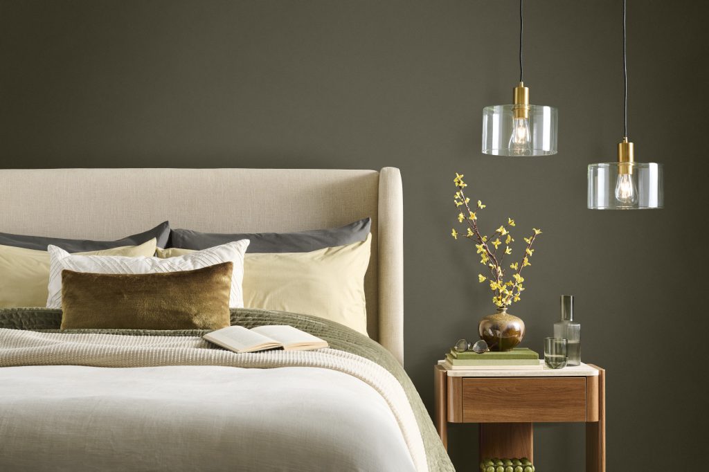 A contemporary and serene bedroom with an accent wall painted in BEHR Grounded, featuring a neutral upholstered bed, layered bedding, a wooden nightstand, and glass pendant lights.