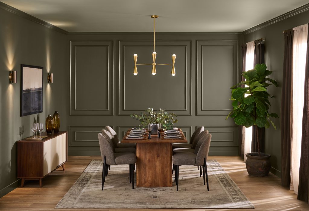 A sophisticated dining room with walls painted in BEHR Grounded, featuring decorative wall molding, a wooden dining table, upholstered chairs, a modern brass chandelier, and warm natural light.