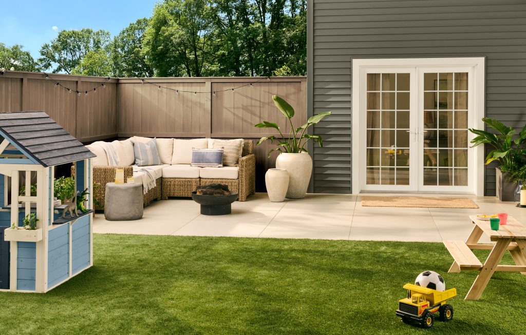 A family-friendly backyard with exterior siding painted in BEHR Grounded, featuring a paved patio, outdoor seating, French doors, a green lawn, and a children’s play area.