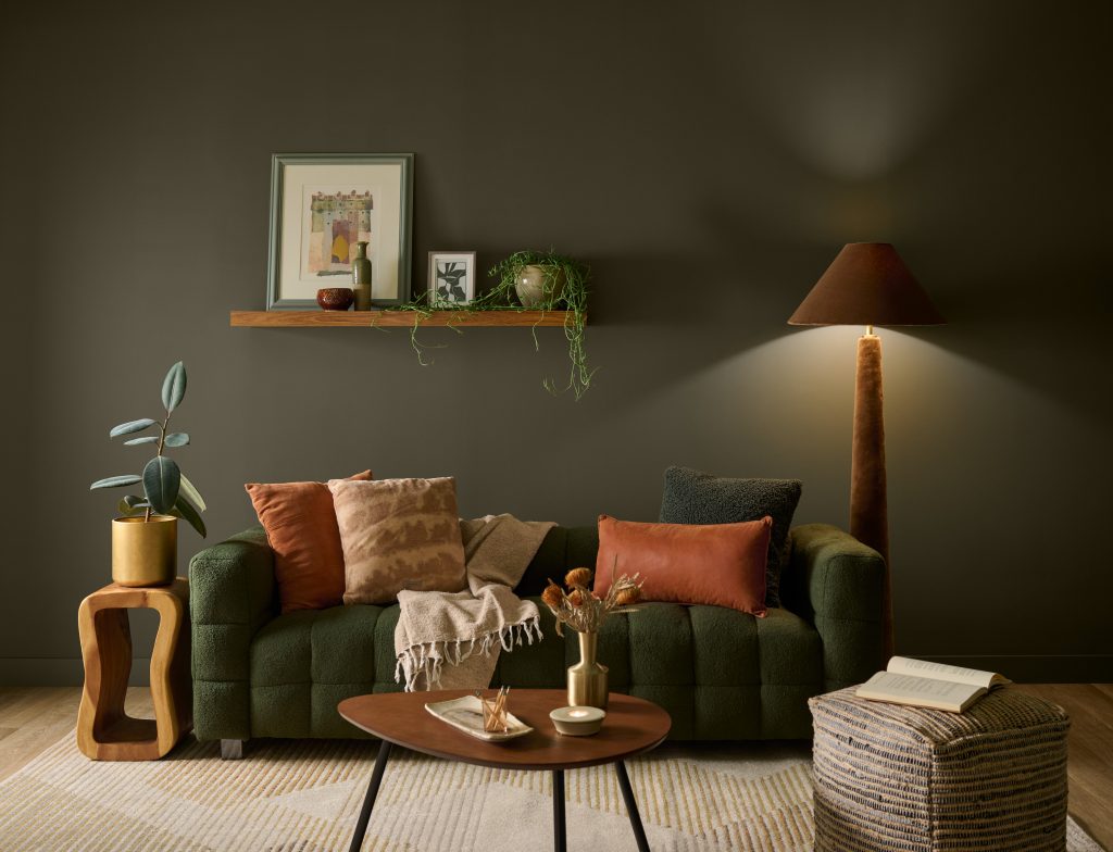 A cozy living room with walls painted in BEHR Grounded, featuring a deep green sofa, warm rust-colored pillows, a wooden coffee table, layered natural textures, and soft ambient lighting.