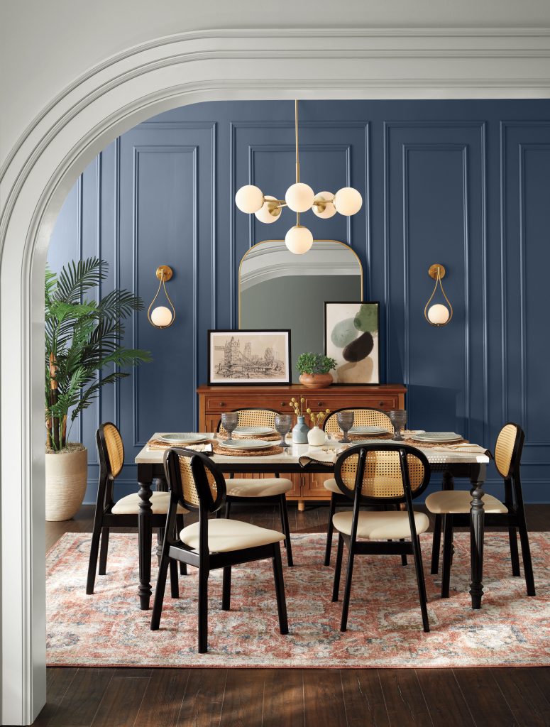 A sophisticated dining room with deep blue paneled walls, a round dining table, black chairs, brass accents, multi-globe chandelier, arched mirror, and layered patterned rug.