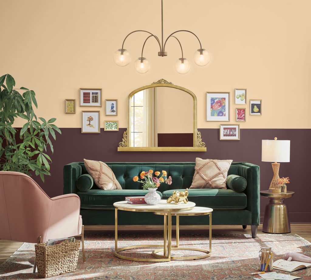 A stylish living room with burgundy lower walls, white upper walls, a green velvet sofa, pink accent chair, gold mirror, gallery wall, arched chandelier, layered rugs, and warm metallic accents.