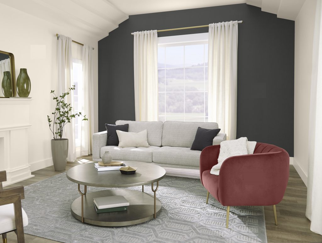 A well-lit living room with a large window, white walls, a charcoal accent wall, very light gray sofa, olive green accent chair, round coffee table, patterned rug, and soft white curtains.