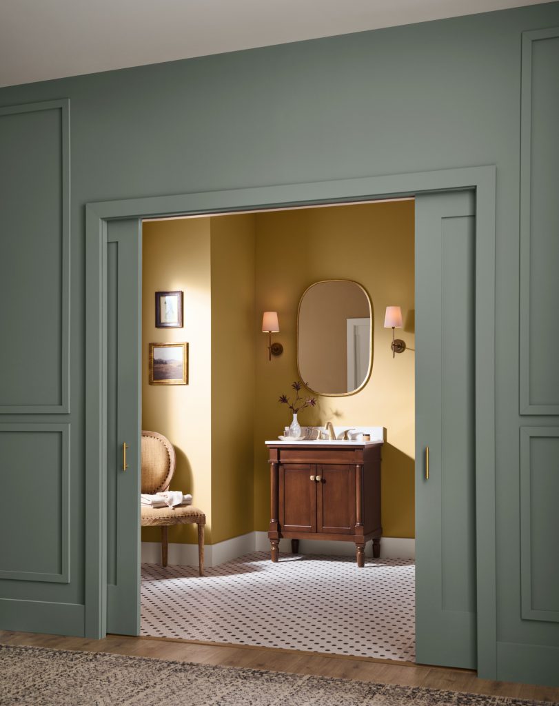 A green-paneled bathroom entryway leading into a warm colored vanity area with a vintage wood cabinet, oval mirror, wall sconces, patterned tile floor, and layered neutral rugs.