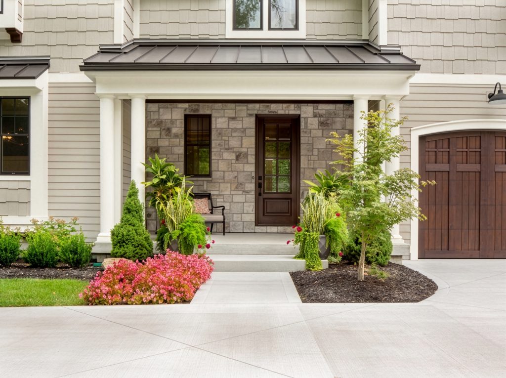 A taupe exterior paint scheme featuring Behr Wheat Bread siding, white trim and columns, stone accents, dark wood doors, and colorful front landscaping.