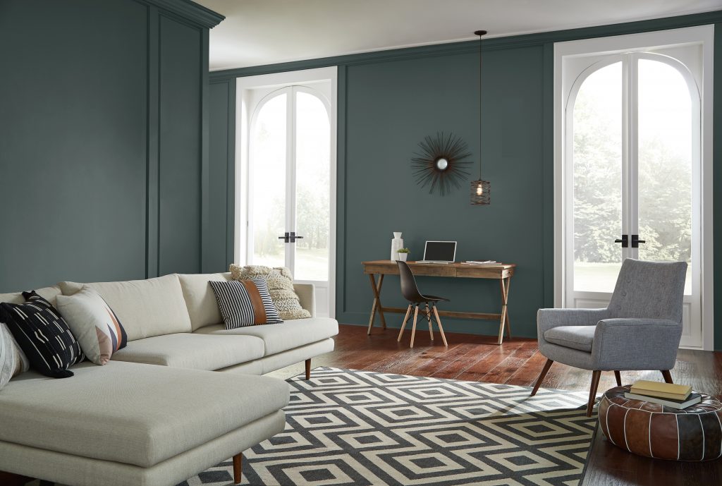 Open living room with dark green painted walls, small desk between tall windows, white sofa, patterned rug, gray chair, and wood flooring.