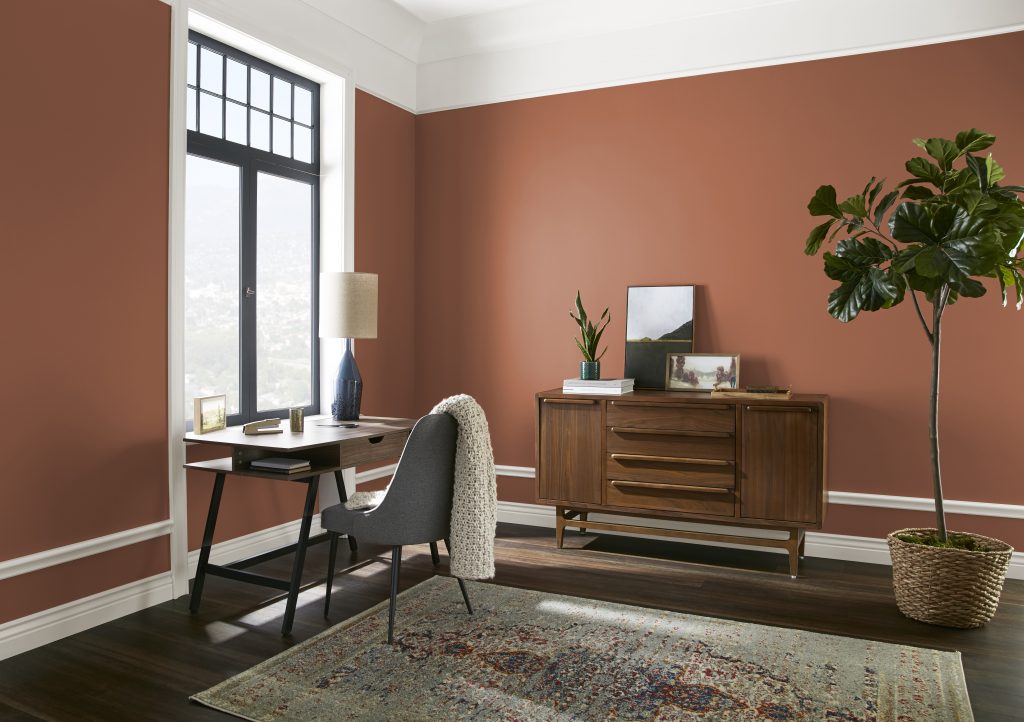 Small home office off a bedroom with an orange-brick tone painted walls, a desk by the window, wood storage cabinet, patterned rug, and indoor plant.