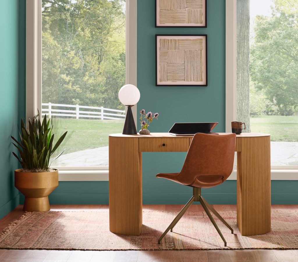 Home office nook painted in smokey jade hue with a small wood desk, windows, framed art, plants, and a chair in an open living area.