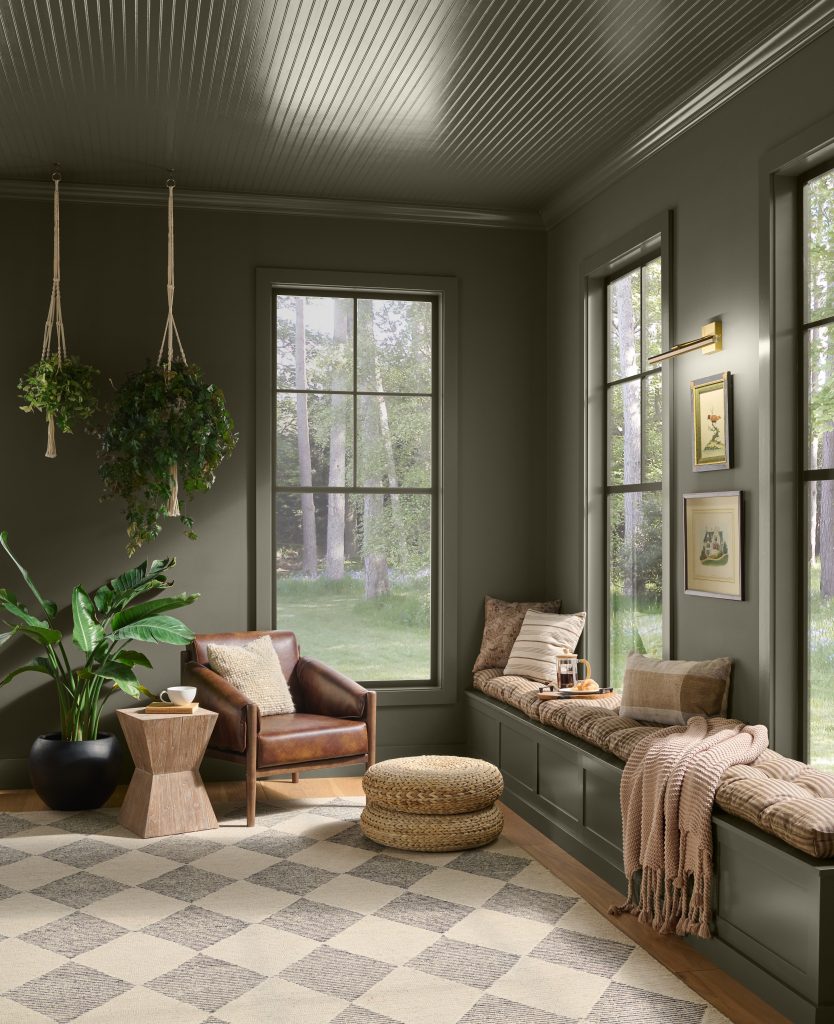 A cozy sunroom painted in BEHR Grounded, featuring a built-in window seat, leather chair, hanging plants, large windows, and a checkered area rug.