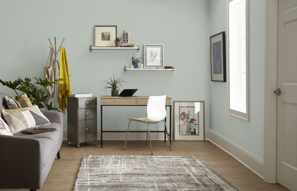 Small living room office with gray-green painted walls, compact desk, white chair, wall shelves, sofa, plants, and light wood flooring.