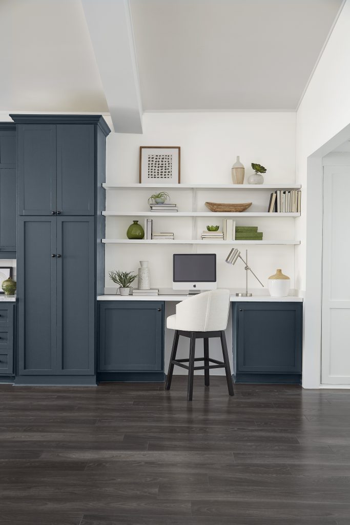 Kitchen-adjacent office area with deep blue-gray cabinetry, built-in desk, open white shelves, stool, computer monitor, and dark wood flooring.
