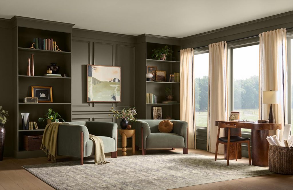 Alt Text: A cozy home office and study with walls painted in BEHR Grounded, featuring built-in bookshelves, two upholstered armchairs, a wooden desk, large windows with cream curtains, and warm natural light.