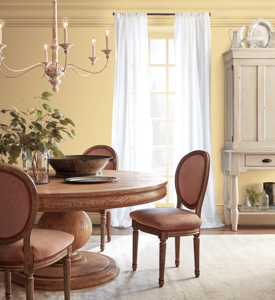 Dining room with Harmonious Gold wall paint, warm wood dining furniture, white curtains, traditional chandelier, and vintage-inspired décor for a romantic Regency-style dining room color palette.