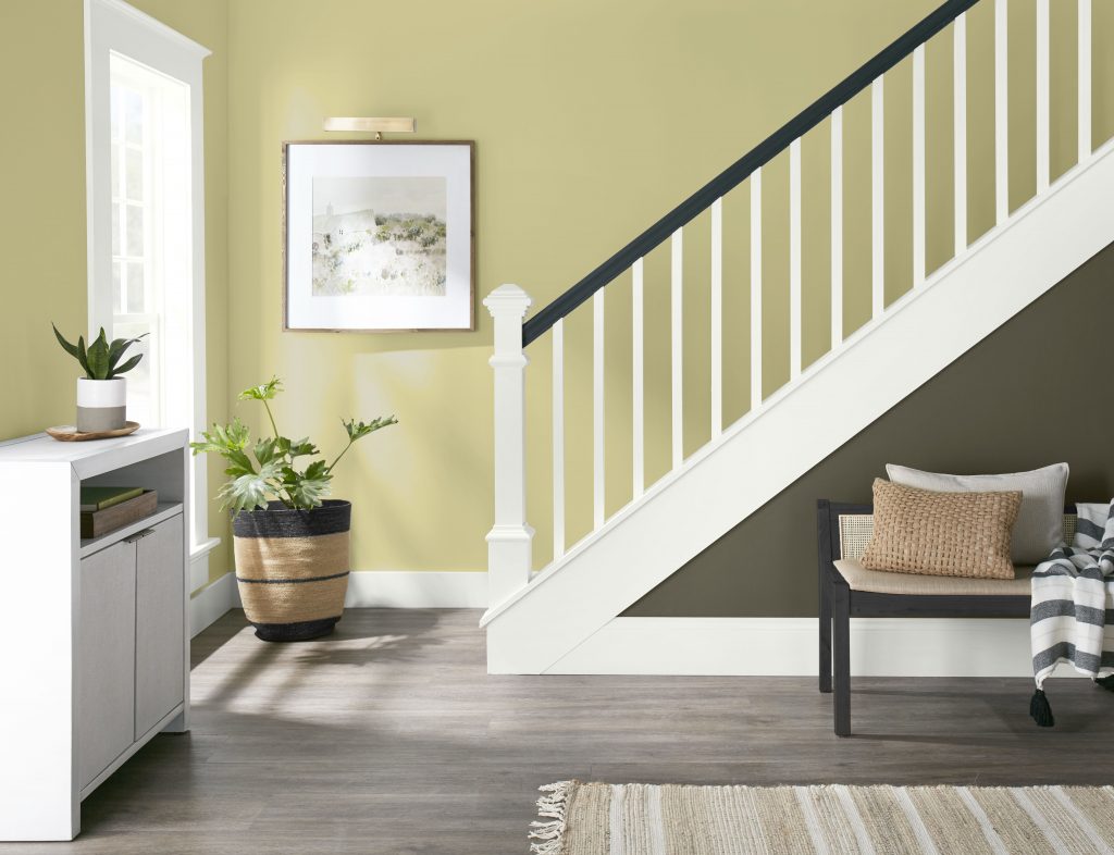 A bright stairway with walls painted BEHR Wheat Grass, crisp Polar Bear trim and railing, and a Grounded accent beneath the stairs. Natural light, greenery, woven textures, and a simple bench create a fresh, welcoming transition space.